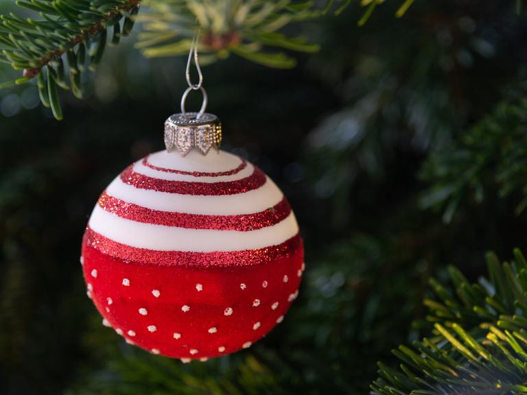 Eine rote Weihnachtskugel mit weißen Punkten hängt an einem geschmückten Weihnachtsbaum.