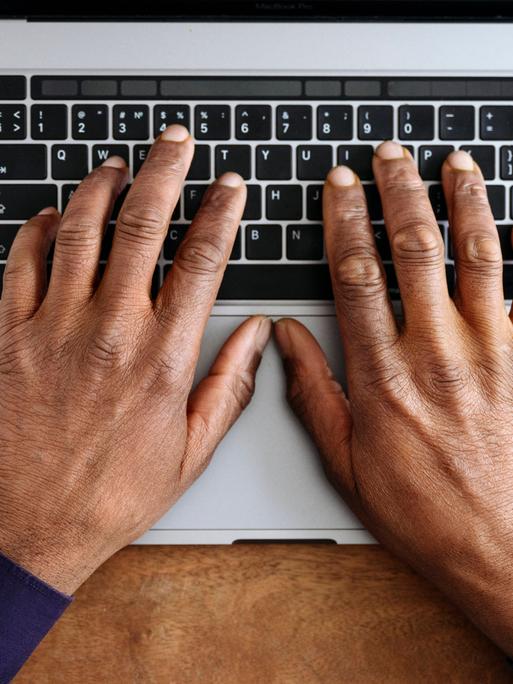 Hände einer schwarzen Person liegen auf der Tastatur eines silberfarbenen Laptops (Symbolfoto).