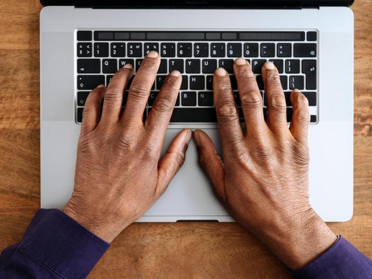 Hände einer schwarzen Person liegen auf der Tastatur eines silberfarbenen Laptops (Symbolfoto).