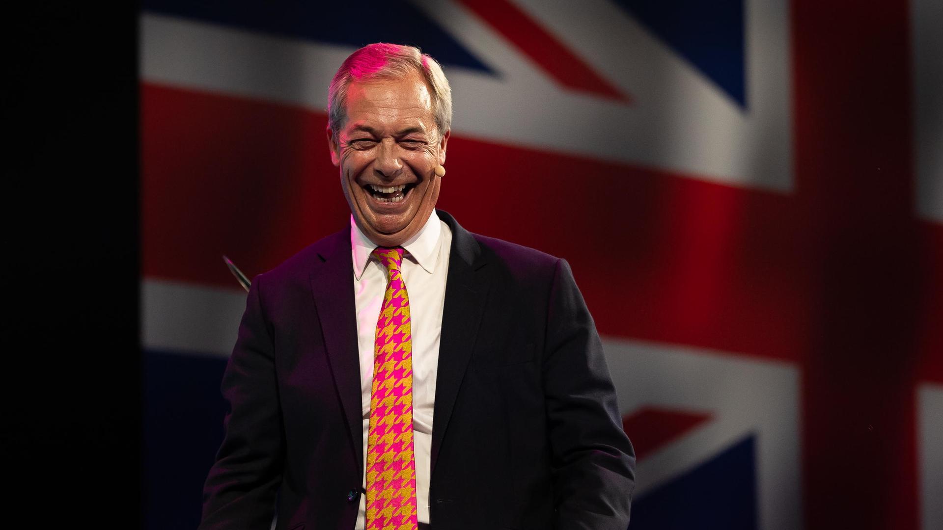 Nigel Farage geht lachend von einer großen Bühne, im Hintergund wird die britische Flagge eingeblendet. 