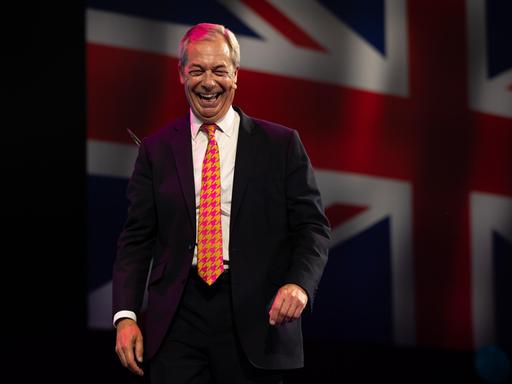 Nigel Farage geht lachend von einer großen Bühne, im Hintergund wird die britische Flagge eingeblendet. 