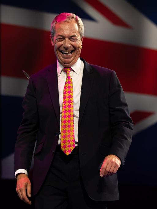 Nigel Farage geht lachend von einer großen Bühne, im Hintergund wird die britische Flagge eingeblendet. 