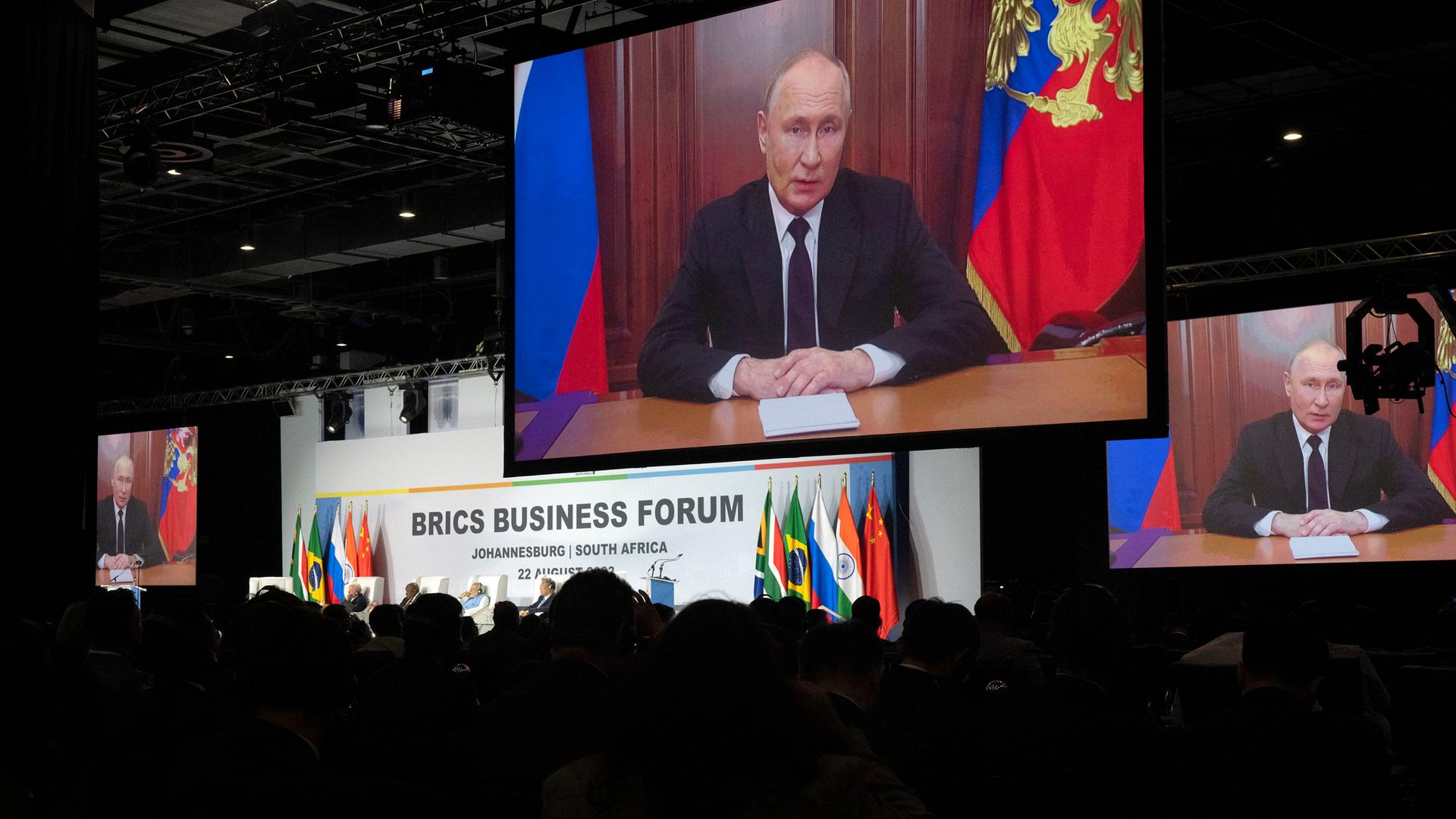 Russlands Präsident Wladimir Putin auf einer großen Videoleinwand in einem Kongresszentrum. Auf der Bühne ist der Schriftzug "Brics Business Forum" zu lesen.