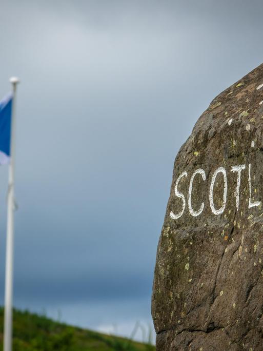 Auf einem Stein steht mit weißer Farbe geschrieben "Schottland". Dahinter weht eine schottische Fahne.