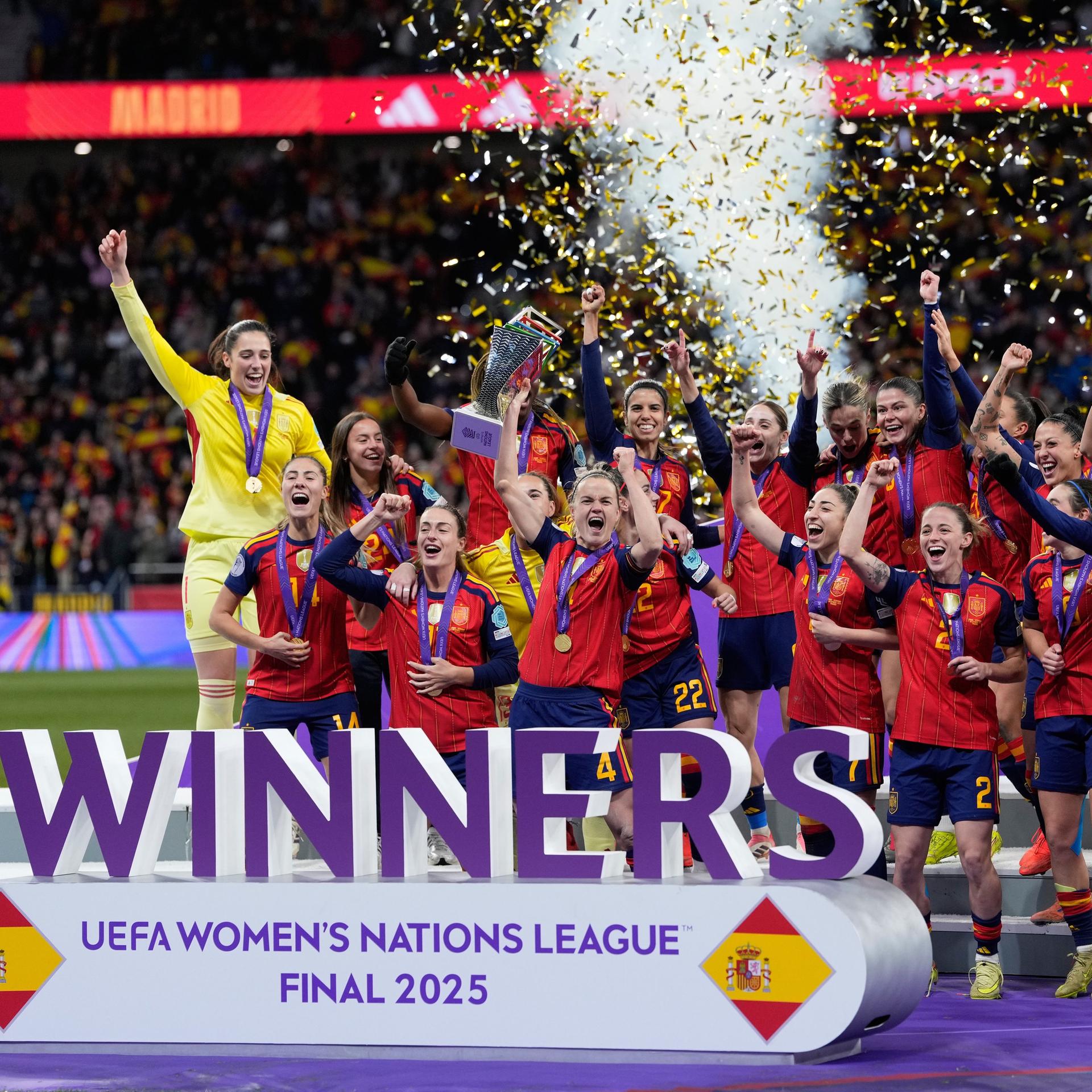 Zu sehen ist das spanische Frauen-Fußball-Nationalteam beim Jubel auf einem Podest. Die Spielerinnen recken ihre Hände hoch und halten den Siegerpokal. Im Hintergund wird Konfeitti in die Luft geblasen.