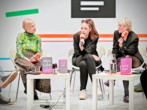 Aiki Mira, Anna-Verena Nosthoff und Veronika Kracher (v.l.n.r.) sitzen mit Dlf-Mikrofonen hinter zwei kleinen Tischen am Deutschlandradio-Stand auf der Leipziger Buchmesse und sprechen miteinander