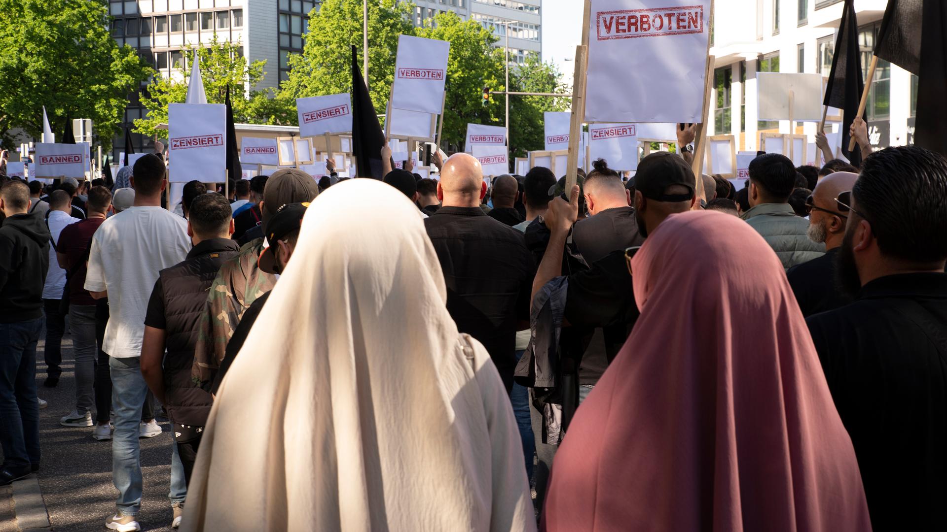 Im Vordergrund des Bildes sieht man zwei Frauen von hinten. Sie tragen Hijab. Im Hintergrund stehen junge Männer auf einer Demonstration. Sie halten Schilder mit den Worten "zensiert" und "verboten" in die Luft.