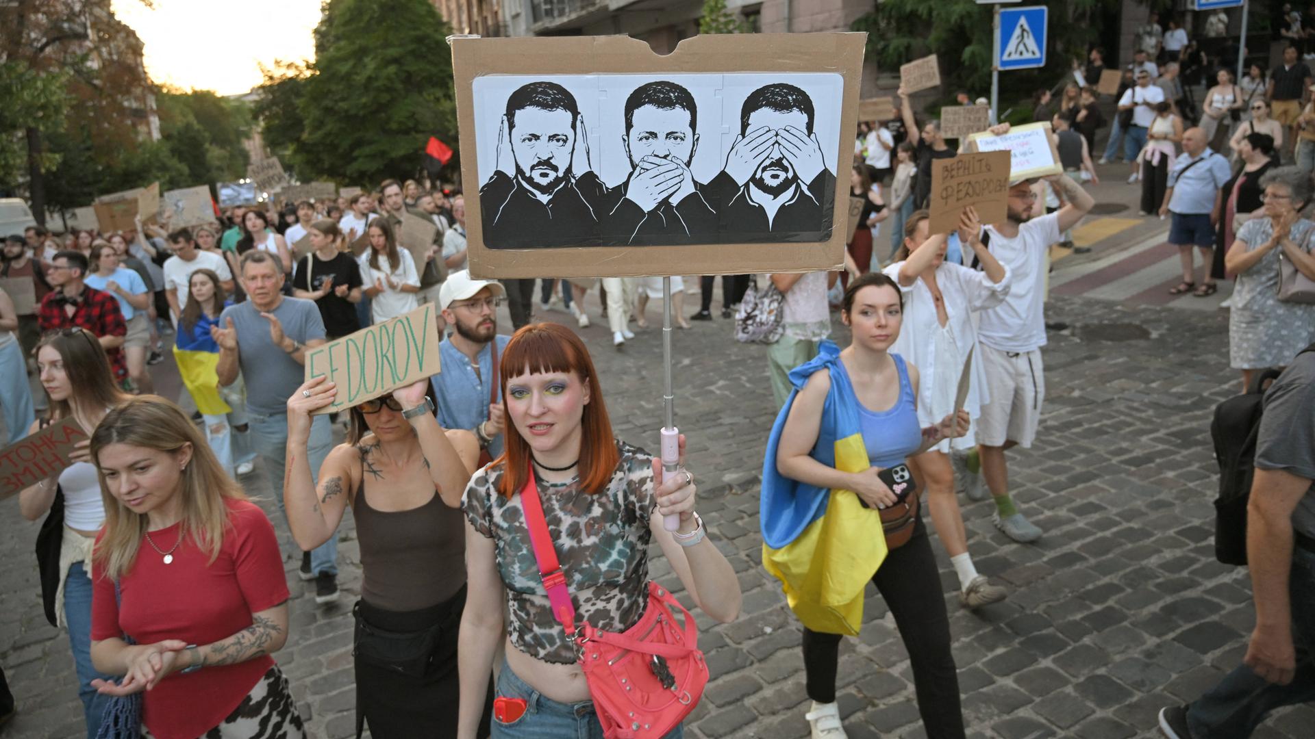 Protestierende in der Ukraine tragen ein Plakat, auf dem drei Zeichnungen von Präsident Selenskyj zu sehen sind. Sie symbolisieren, dass er nichts sehe, nichts hören und nichts sagen wolle.