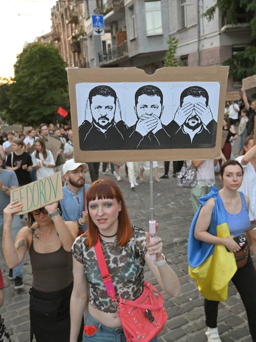 Protestierende in der Ukraine tragen ein Plakat, auf dem drei Zeichnungen von Präsident Selenskyj zu sehen sind. Sie symbolisieren, dass er nichts sehe, nichts hören und nichts sagen wolle.