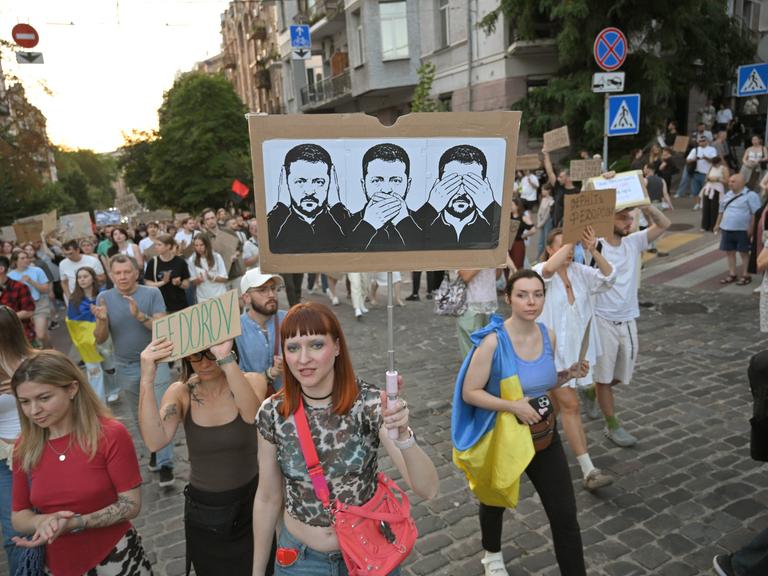 Protestierende in der Ukraine tragen ein Plakat, auf dem drei Zeichnungen von Präsident Selenskyj zu sehen sind. Sie symbolisieren, dass er nichts sehe, nichts hören und nichts sagen wolle.
