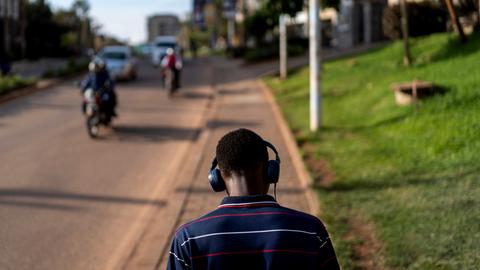 Rückansicht eines jungen Mannes, der auf einem Gehsteig in Kampala spazieren geht. Er trägt Kopfhörer. Auf der Straße kommt ihm ein Motorrad entgegen Rückansicht eines jungen Mannes, der auf einem Gehsteig in Kampala spazieren geht. Er trägt Kopfhörer. Auf der Straße kommt ihm ein Motorrad entgegen