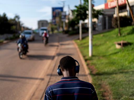 Rückansicht eines jungen Mannes, der auf einem Gehsteig in Kampala spazieren geht. Er trägt Kopfhörer. Auf der Straße kommt ihm ein Motorrad entgegen