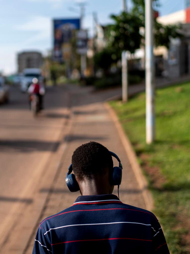 Rückansicht eines jungen Mannes, der auf einem Gehsteig in Kampala spazieren geht. Er trägt Kopfhörer. Auf der Straße kommt ihm ein Motorrad entgegen