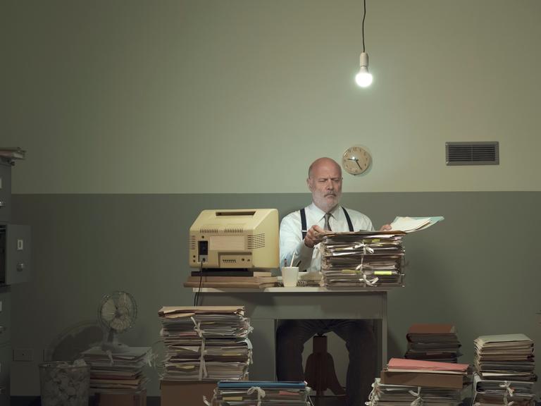 Symbolbild: Frustrierter Geschäftsmann im Vintage-Stil, der in einem heruntergekommenen Büro arbeitet und mit Papierkram überfordert ist.