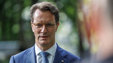 Ministerpräsident Hendrik Wüst steht im Freien, im Hintergrund Bäume. Er trägt einen hellblauen Sakko mit NRW-Anstecker am Revers und eine hellbraune Brille.