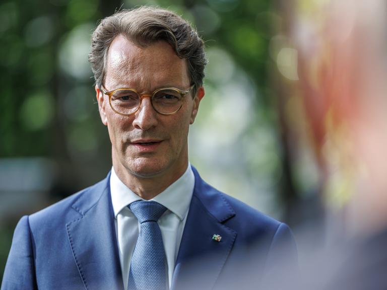 Ministerpräsident Hendrik Wüst steht im Freien, im Hintergrund Bäume. Er trägt einen hellblauen Sakko mit NRW-Anstecker am Revers und eine hellbraune Brille. Ministerpräsident Hendrik Wüst steht im Freien, im Hintergrund Bäume. Er trägt einen hellblauen Sakko mit NRW-Anstecker am Revers und eine hellbraune Brille.