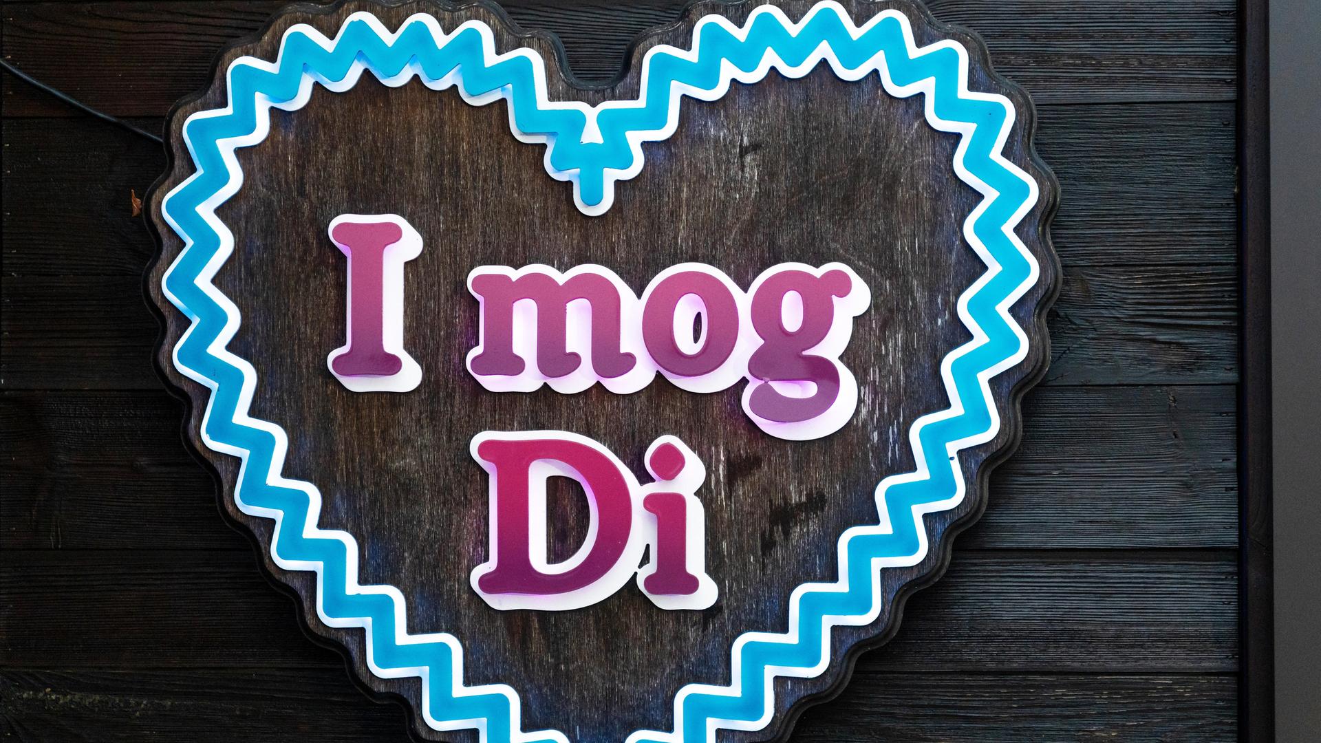 Ein Schild in Herzform zeigt den Schriftzug "I mog Di" (bairisch für "Ich hab' dich lieb"). Das blaue Herz erinnert an ein Lebkuchenherz.