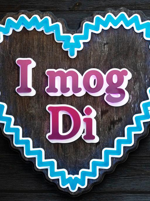 Ein Schild in Herzform zeigt den Schriftzug "I mog Di" (bairisch für "Ich hab' dich lieb"). Das blaue Herz erinnert an ein Lebkuchenherz.