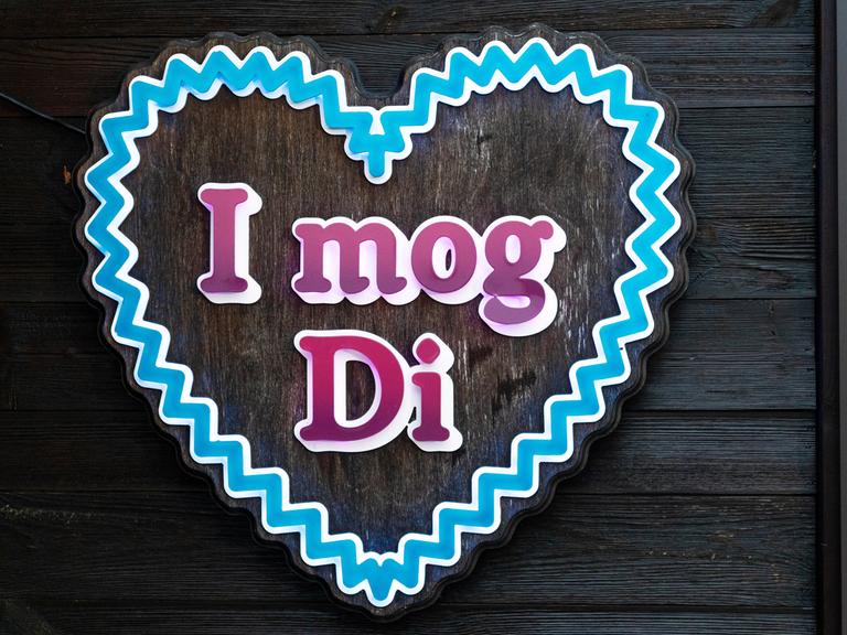 Ein Schild in Herzform zeigt den Schriftzug "I mog Di" (bairisch für "Ich hab' dich lieb"). Das blaue Herz erinnert an ein Lebkuchenherz.