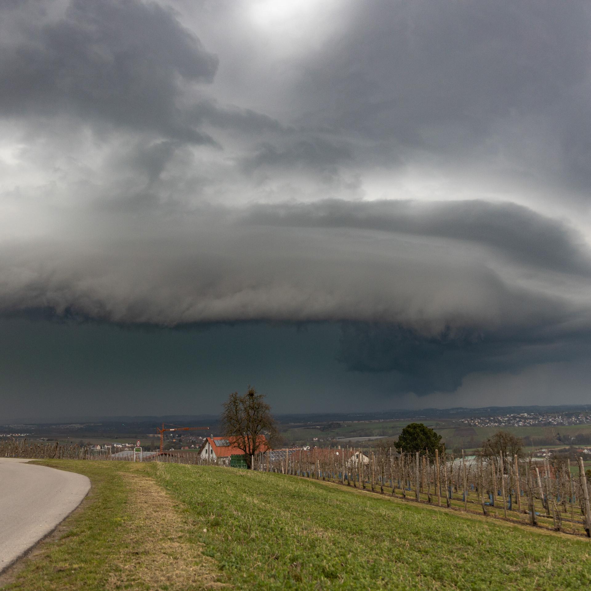 Über einer Ortschaft in einer grünen Landschaft braut sich am Himmel eine dunkle Gewitterzelle zusammen. 