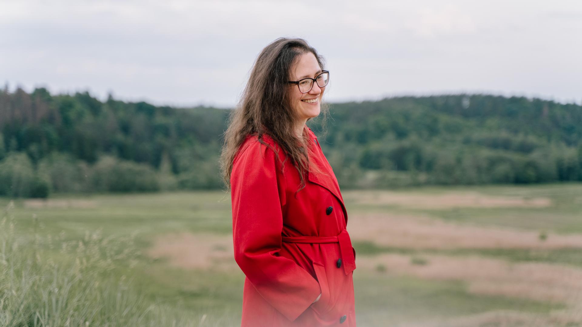 Eine Frau mit langen Haaren und Brille steht in einem roten Mantel mitten in einer Landschaft