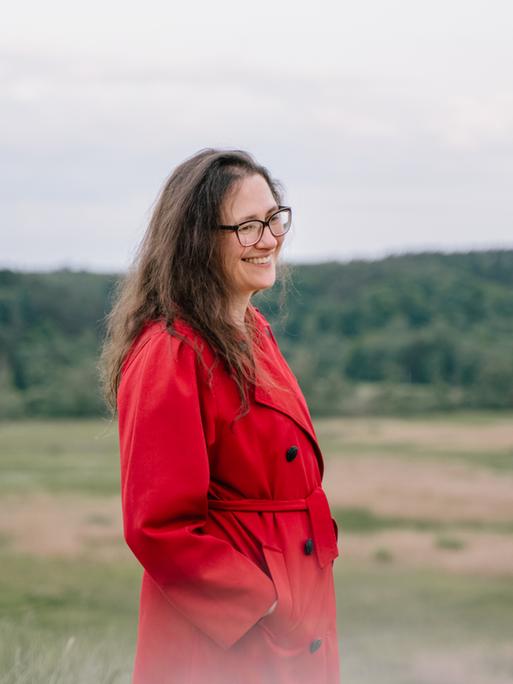 Eine Frau mit langen Haaren und Brille steht in einem roten Mantel mitten in einer Landschaft