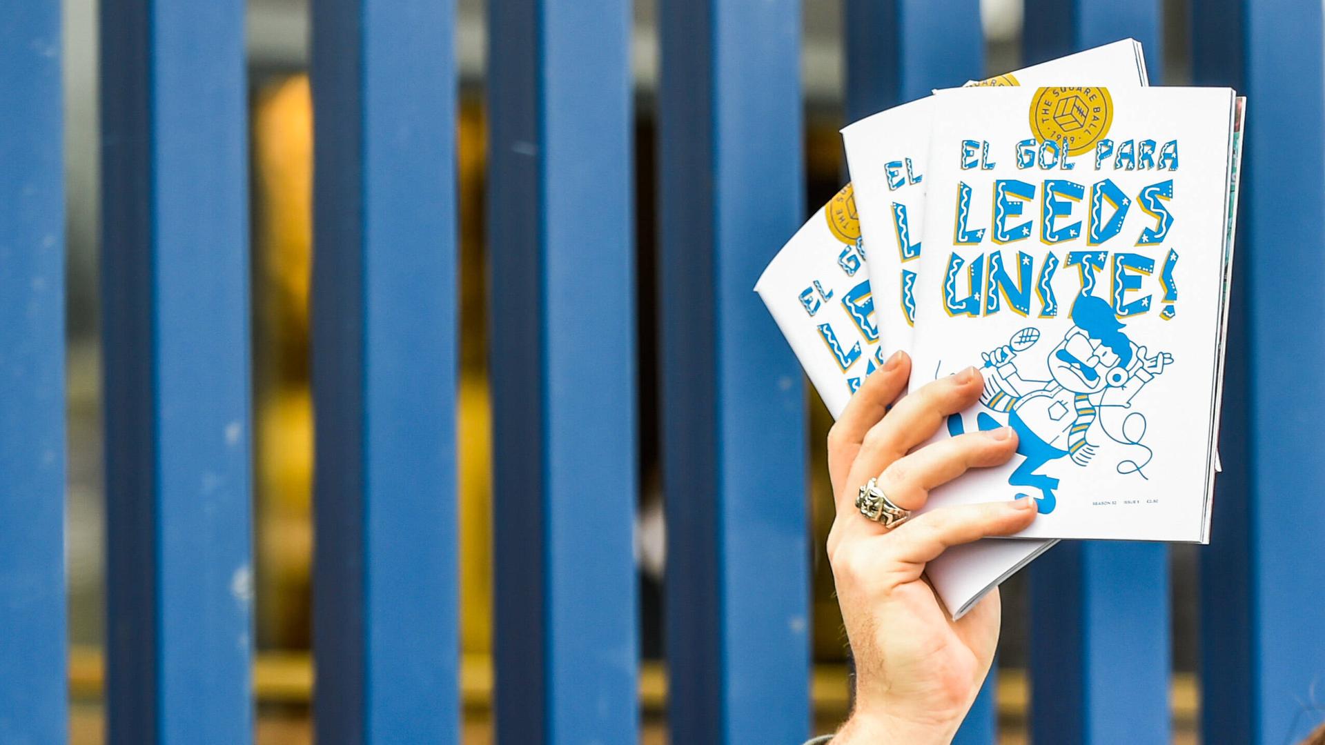 Eine männliche Hand mit einem Ring hält drei Exemplare des Fanzines "El Gol Para Leeds Unite!" hoch, im Hintergrund sind blaue Zaunlatten zu sehen. 