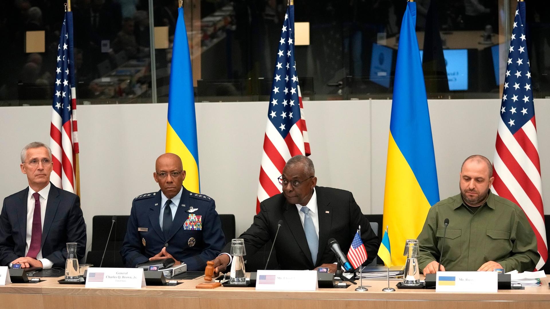 Die vier sitzen nebeneinander am langen Tisch vor Flaggen der USA und der Ukraine. Austin eröffnet die Sitzung mit einem Holzhammmer. 