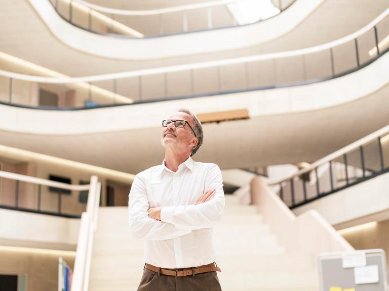 Ein Mann mit grauen Haaren steht in einem modernen Gebäude mit verschränkten Armen und blickt nach oben. Im Hintergrund sind mehrere offene Stockwerke und Treppen zu sehen.