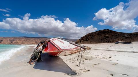 An einem Strand der jemenitischen Insel Sokotra schieben Fischende ein Boot auf den hellen Sandstrand, vor blauem Himmel.