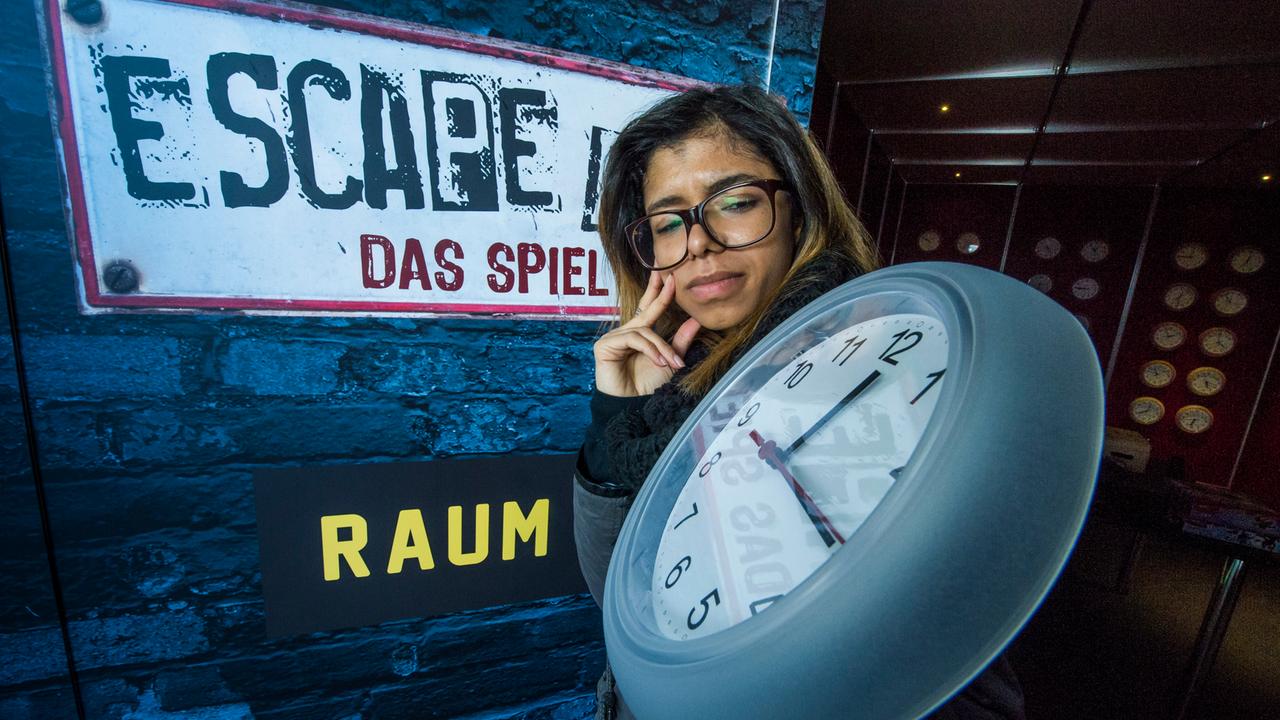 Eine Frau hält eine Wanduhr in der Hand. Dahinter steckt der Schriftzug "Escape Room - das Spiel".