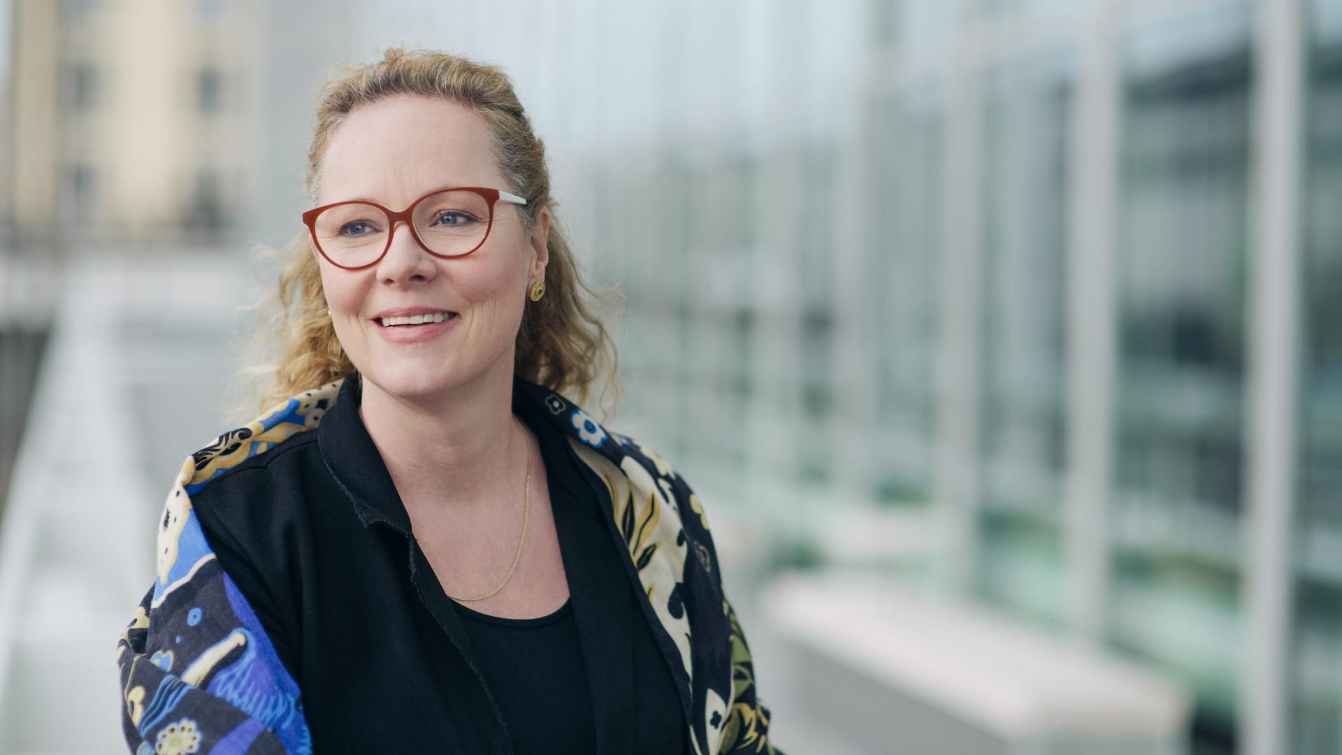 Eine Frau mit blondem Haar steht vor einer Glasfassade. Sie trägt eine dunkelrote Brille und blickt lächelnd links aus dem Bild heraus. Sie trägt ein schwarzes Oberteil und eine geblümte Jacke darüber. Eine Frau mit blondem Haar steht vor einer Glasfassade. Sie trägt eine dunkelrote Brille und blickt lächelnd links aus dem Bild heraus. Sie trägt ein schwarzes Oberteil und eine geblümte Jacke darüber.