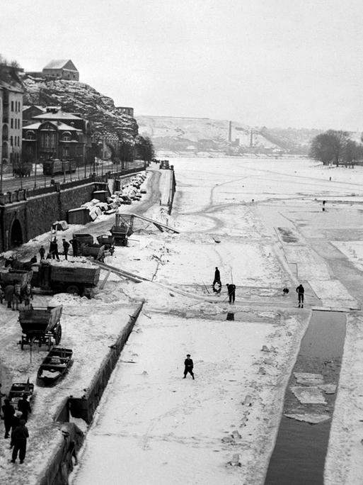 Ein historisches Bild zeigt eine winterliche Flusslandschaft am Rande einer Stadt.