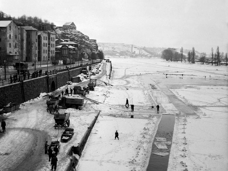 Ein historisches Bild zeigt eine winterliche Flusslandschaft am Rande einer Stadt.