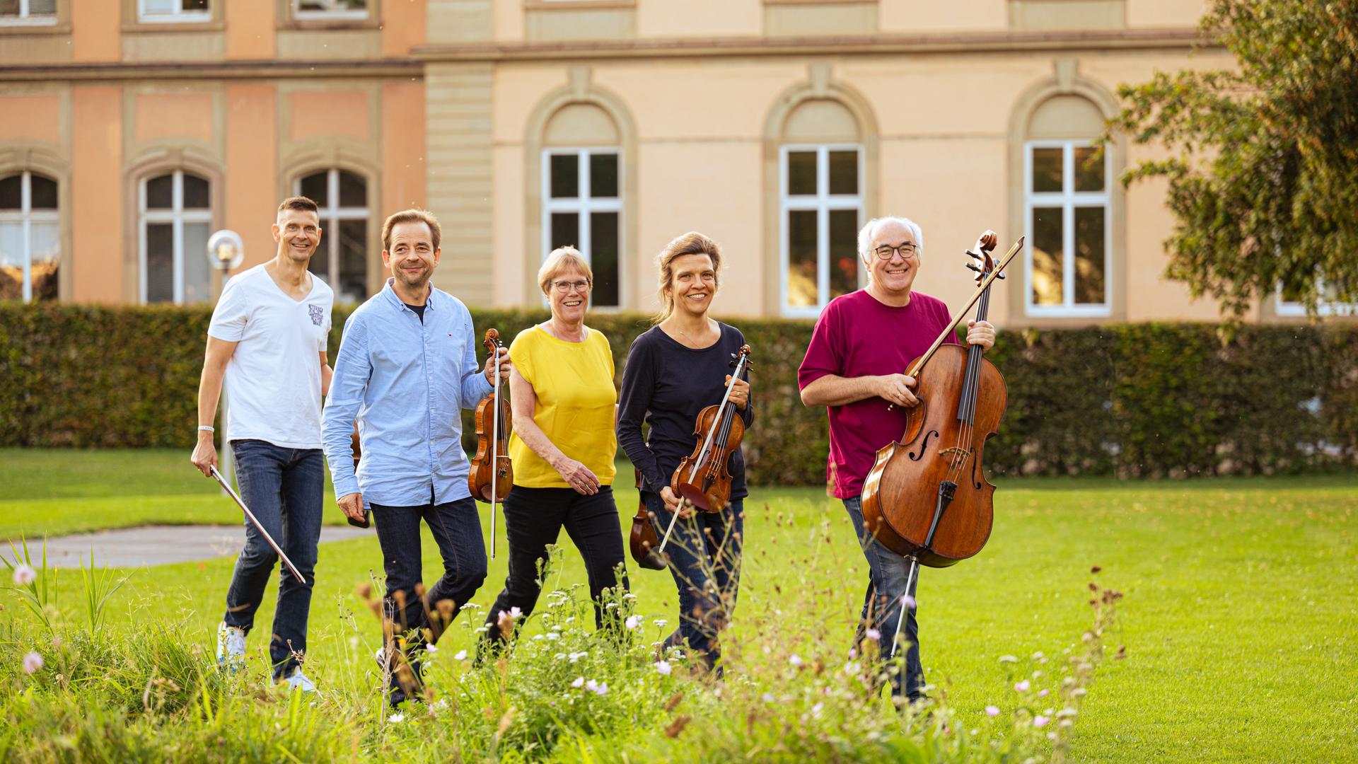 Ein Streichquintett mit einfarbigen Oberteilen läuft mit Instrumenten über eine Wiese. Es sind drei Männer und zwei Frauen, alle lachen. Im Hintergrund sind Häuserfassaden zu sehen.