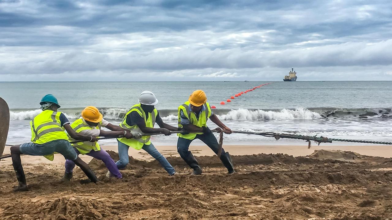 Einige Männer ziehen am Strand an einem Kabel. Im Hintergrund sind das Meer und ein Schiff zu sehen. 