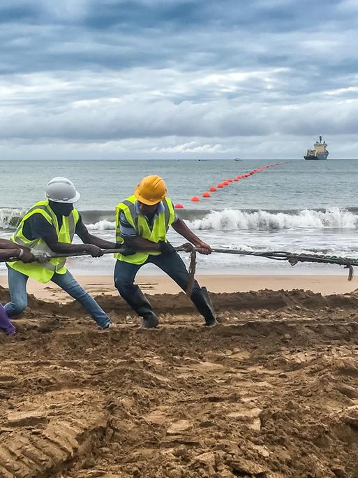 Einige Männer ziehen am Strand an einem Kabel. Im Hintergrund sind das Meer und ein Schiff zu sehen. 