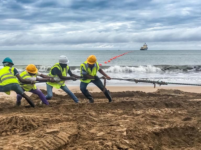 Einige Männer ziehen am Strand an einem Kabel. Im Hintergrund sind das Meer und ein Schiff zu sehen. 