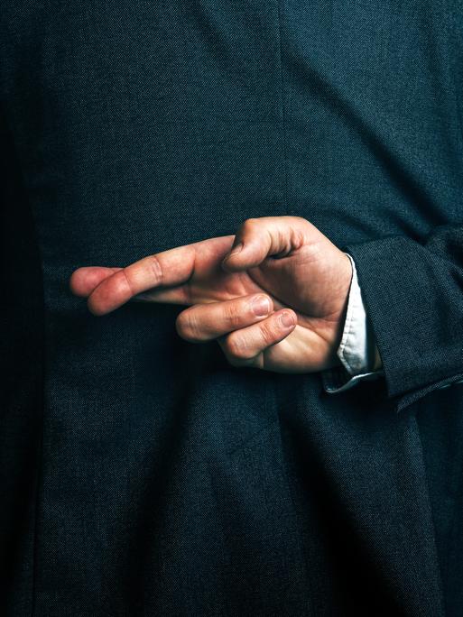 Eine Person im Anzug hält hinter dem Rücken die Finger einer Hand überkreuzt.