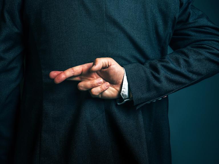 Eine Person im Anzug hält hinter dem Rücken die Finger einer Hand überkreuzt.