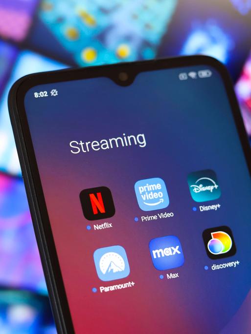 Ein Smartphone mit den Apps der Streaming-Anbieter Amazon, Disney und Netflix vor dem Hintergrund anderer verschwommener Bildschirme