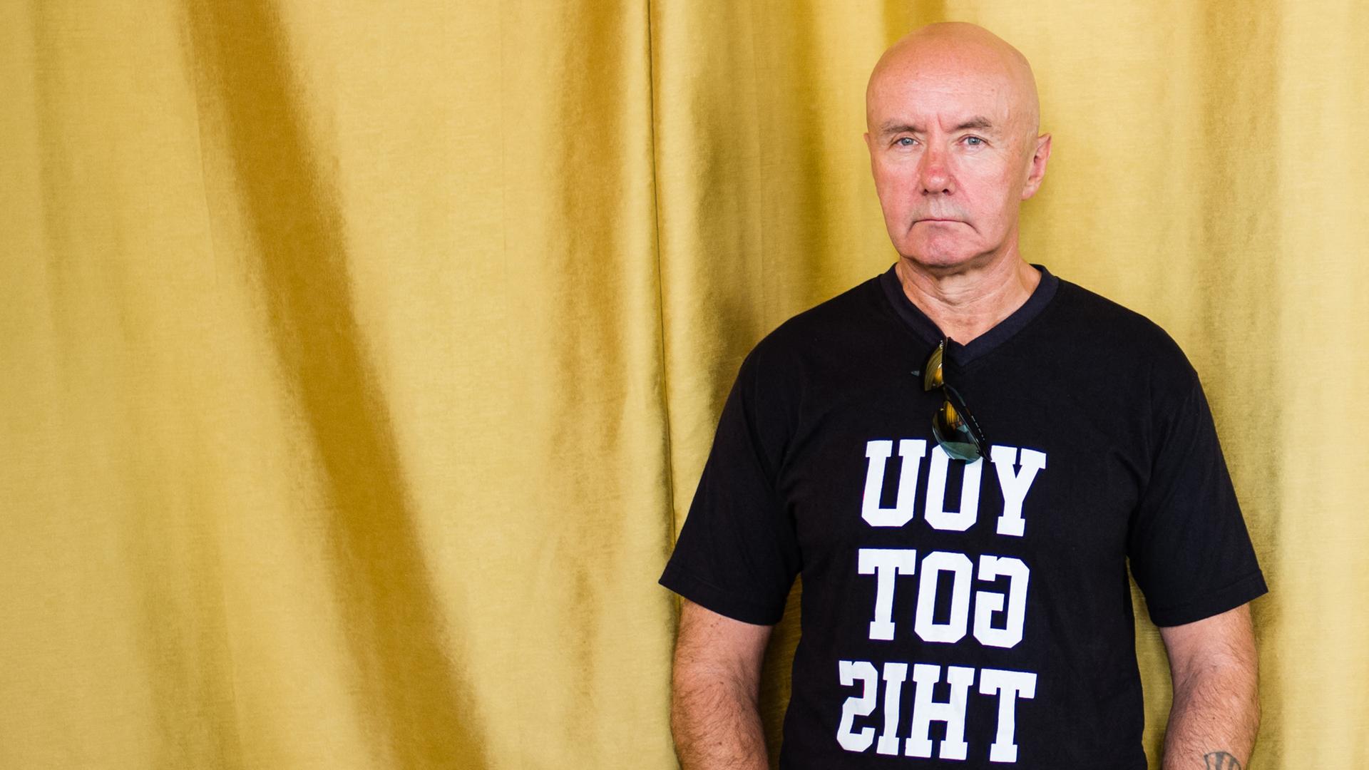Porträt des schottischen Schriftstellers Irvine Welsh, er trät ein schwarzes T-Shirt mit der spiegelverkehrten Aufschrift "YOU GOT THIS" und steht vor einem goldenen Vorhang. Porträt des schottischen Schriftstellers Irvine Welsh, er trät ein schwarzes T-Shirt mit der spiegelverkehrten Aufschrift "YOU GOT THIS" und steht vor einem goldenen Vorhang.