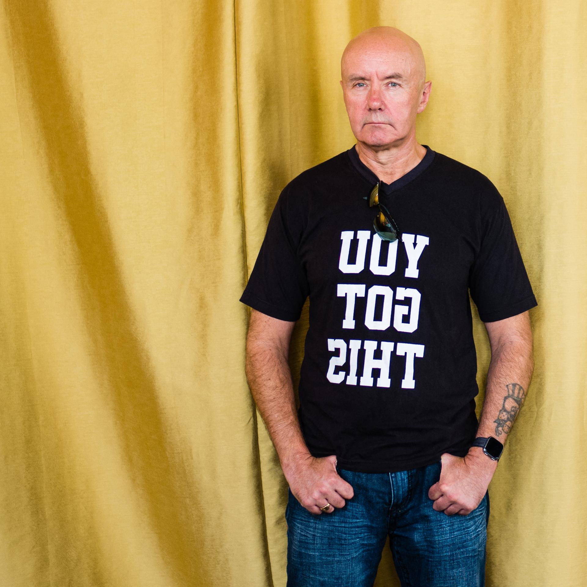 Porträt des schottischen Schriftstellers Irvine Welsh, er trät ein schwarzes T-Shirt mit der spiegelverkehrten Aufschrift "YOU GOT THIS" und steht vor einem goldenen Vorhang.
