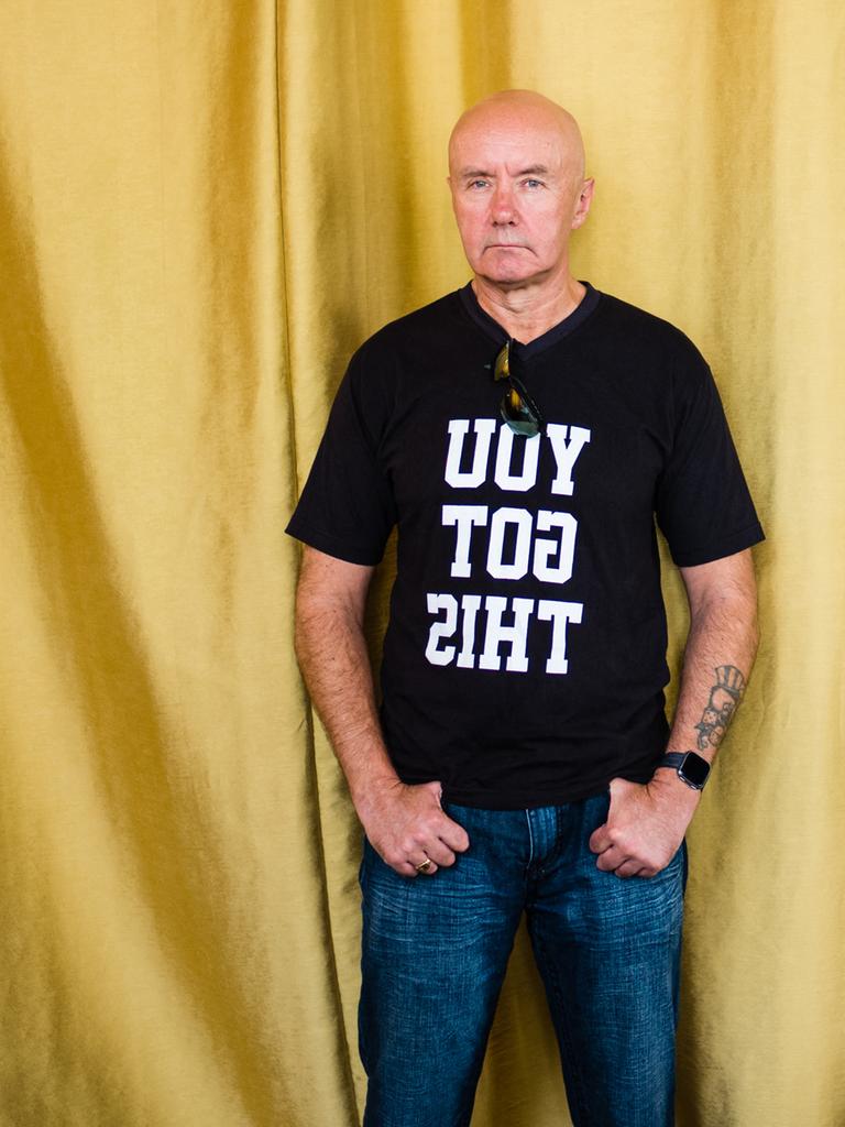 Porträt des schottischen Schriftstellers Irvine Welsh, er trät ein schwarzes T-Shirt mit der spiegelverkehrten Aufschrift "YOU GOT THIS" und steht vor einem goldenen Vorhang.