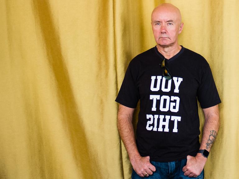 Porträt des schottischen Schriftstellers Irvine Welsh, er trät ein schwarzes T-Shirt mit der spiegelverkehrten Aufschrift "YOU GOT THIS" und steht vor einem goldenen Vorhang. Porträt des schottischen Schriftstellers Irvine Welsh, er trät ein schwarzes T-Shirt mit der spiegelverkehrten Aufschrift "YOU GOT THIS" und steht vor einem goldenen Vorhang.