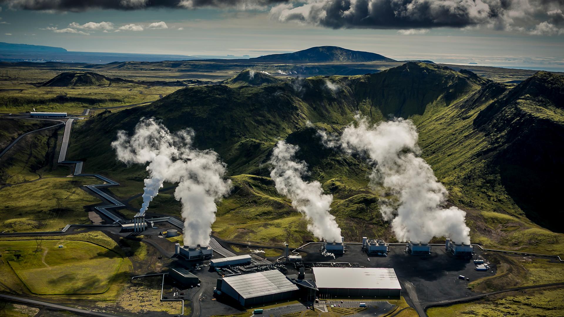 Luftaufnahme eines geothemalen Kraftwerks in Island, aus dessen Schornsteinen weißer Dampf aufsteigt.
