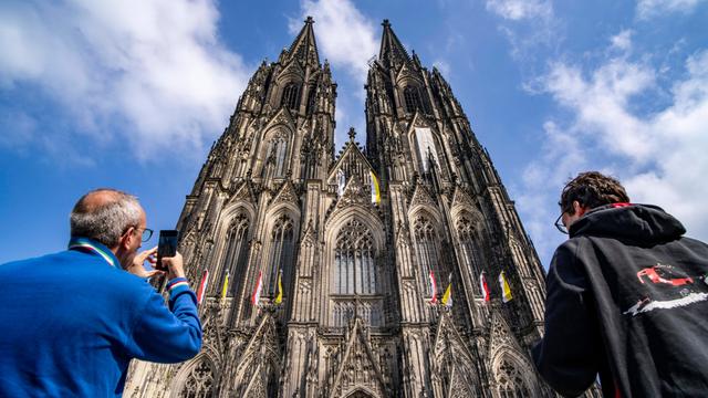 2 Personen stehen vor dem Kölner Dom. Sie sind von hinten zu sehen. Eine Person fotografiert den Kölner Dom.