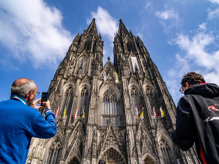 Zwei Personen stehen vor dem Kölner Dom. Sie sind von hinten zu sehen. Eine Person fotografiert den Kölner Dom.