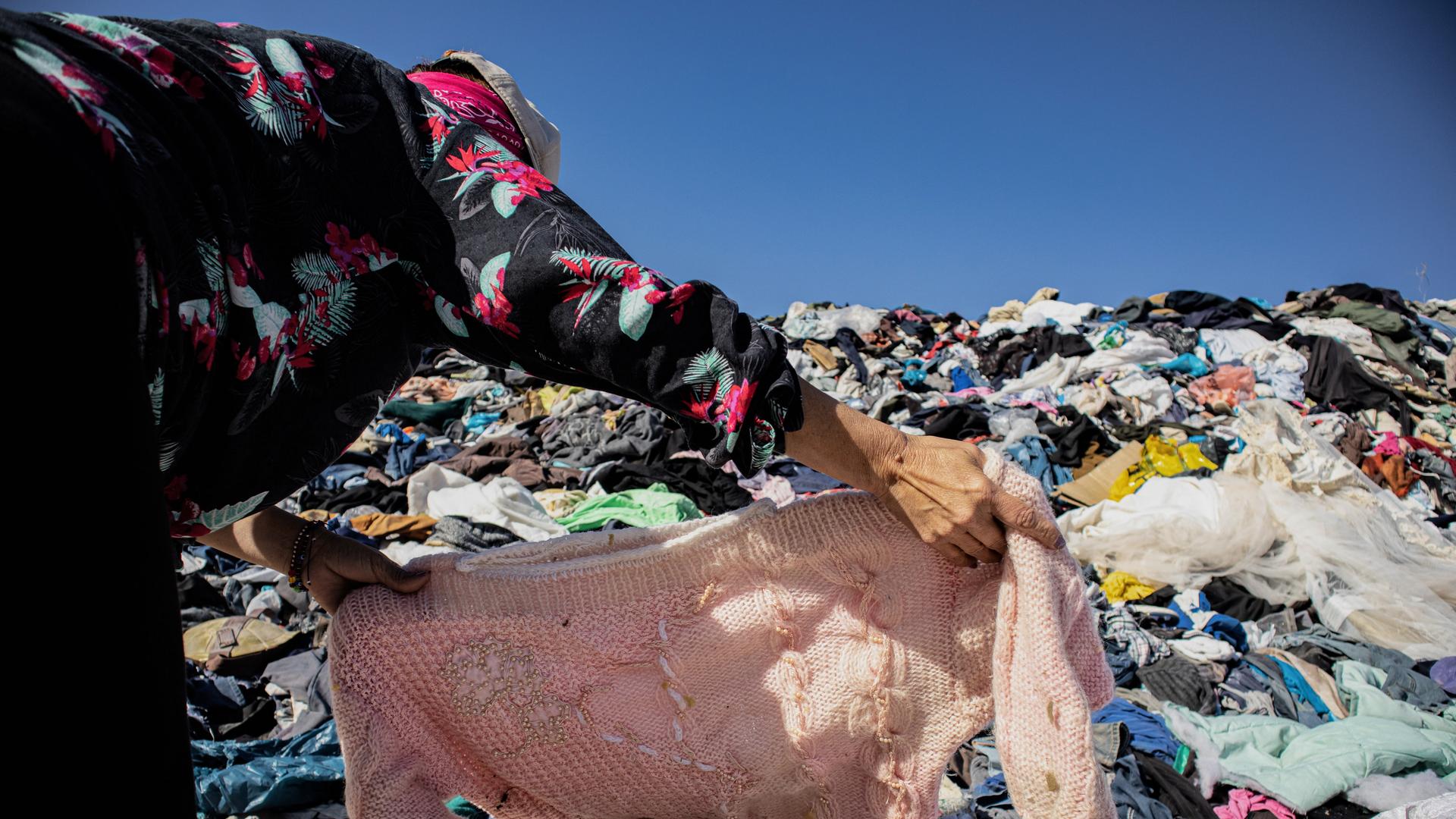 Eine Frau sucht unter einem Berg gebrauchter Kleidern in einer Müll-Deponie ein Kleidungsstück. In der nahe gelegenen Freihandelszone von Iquique kamen 2021 bis Oktober 29.178 Tonnen gebrauchter Kleidung an.