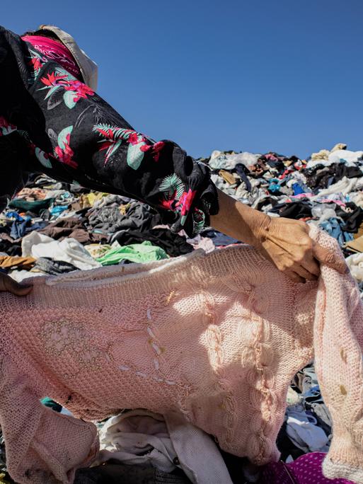 Eine Frau sucht unter einem Berg gebrauchter Kleidern in einer Müll-Deponie ein Kleidungsstück. In der nahe gelegenen Freihandelszone von Iquique kamen 2021 bis Oktober 29.178 Tonnen gebrauchter Kleidung an.